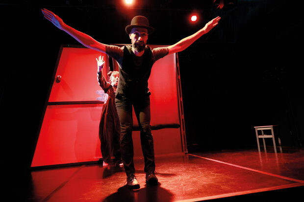 Ein Schauspieler mit schwarzem Hut steht auf einer rot ausgeleuchteten Bühne, die Arme weit ausgebreitet. Im Hintergrund ist eine Schauspielerin zu sehen, die ebenfalls ihre Arme hebt.