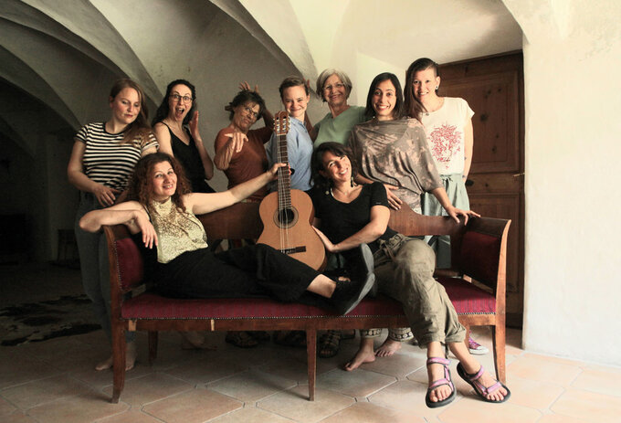 Gruppenfoto von neun Frauen des Chors, aufgenommen in einem hellen Raum mit Gewölbedecke. Zwei sitzen auf einer Holzbank mit Gitarre, die anderen stehen dahinter und zeigen fröhliche, lebendige Posen.