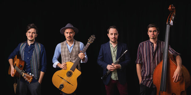 Vier Musiker stehen nebeneinander vor schwarzem Hintergrund. Zwei halten Gitarren, einer eine Klarinette und einer einen Kontrabass.