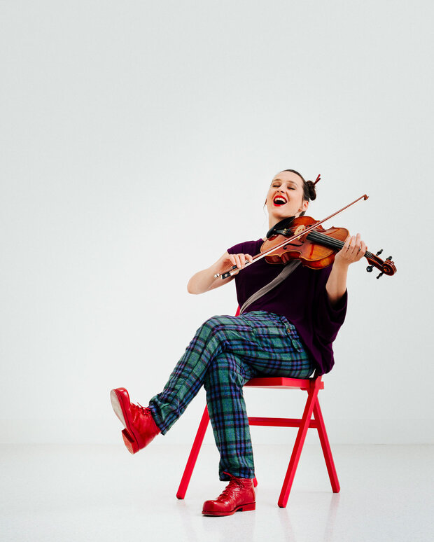 Eine Frau sitzt auf einem roten Stuhl vor weißem Hintergrund und spielt Geige. Sie trägt karierte Hosen und rote Schuhe. Beim Spielen lächelt sie und wirkt fröhlich und energiegeladen.