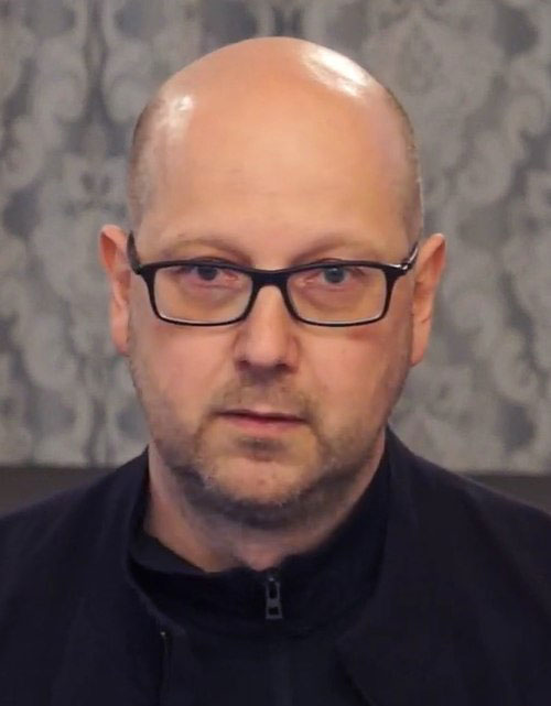 Porträt eines Mannes mit Brille und Glatze, der ruhig in die Kamera blickt. Der Hintergrund zeigt ein graues, gemustertes Muster.