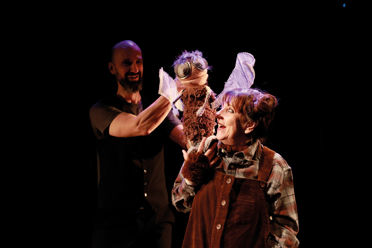 Eine Schauspielerin und ein Puppenspieler auf der Bühne. Der Puppenspieler hält eine große Fliegenpuppe mit ausgebreiteten Flügeln, während die Schauspielerin lachend zu ihr aufschaut.