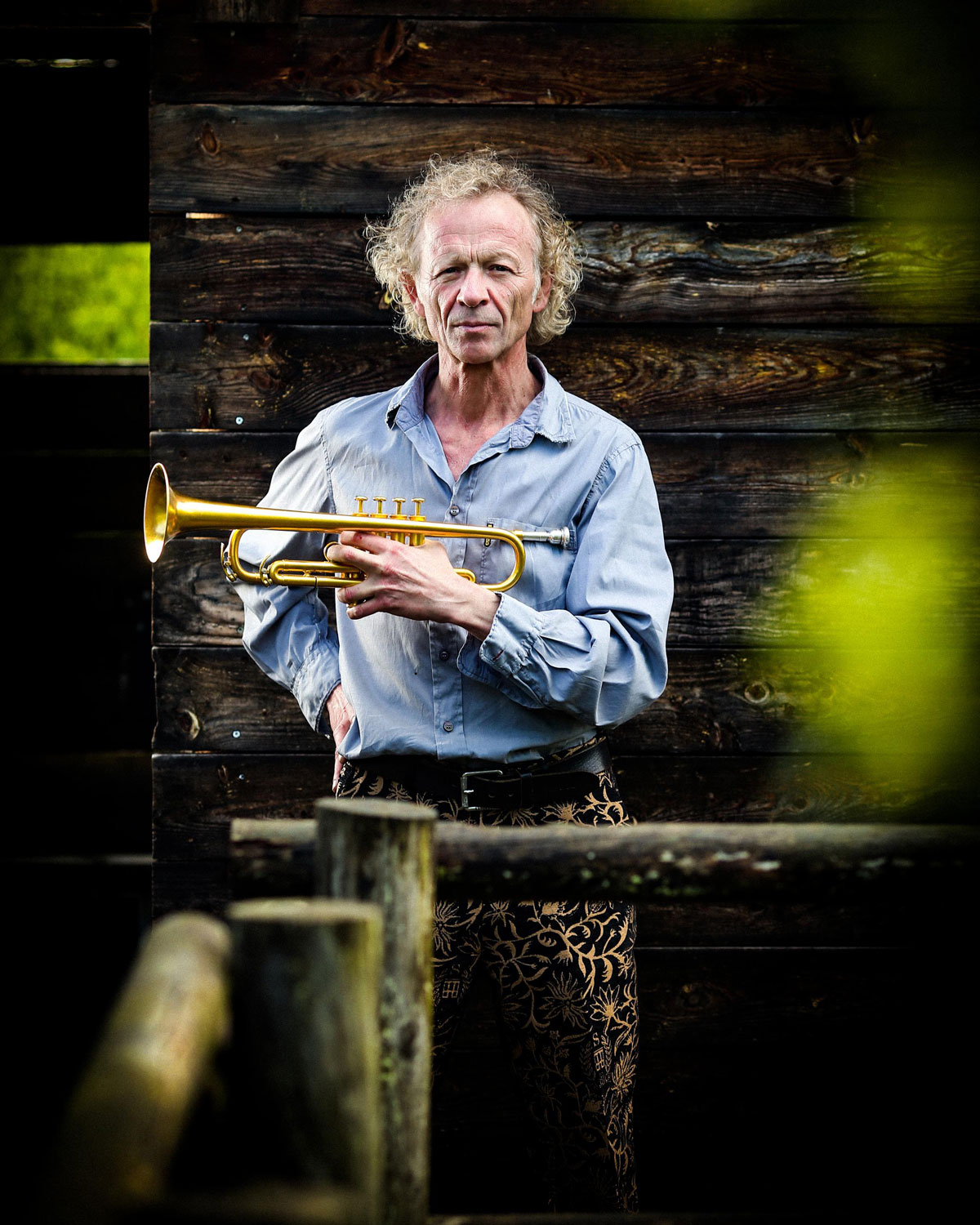 Ein Mann mit lockigem, grauem Haar steht vor einer dunklen Holzwand und hält eine Trompete in der Hand. Er schaut ruhig zur Seite. Im Vordergrund ist unscharfes grünes Laub zu sehen.